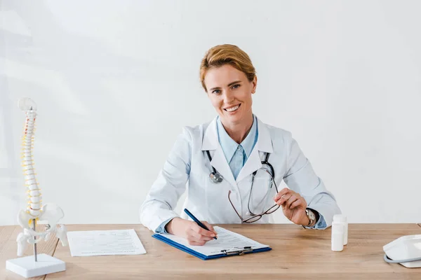 Happy doctor holding glasses and pen near clipboard in clinic — Stock Photo
