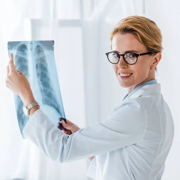 Schöne und fröhliche Ärztin mit Brille, die in die Kamera schaut und Röntgenaufnahmen in der Klinik macht — Stockfoto