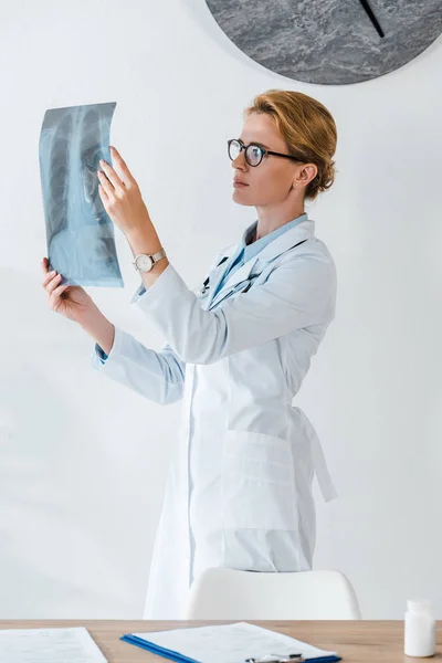 Belle dans les lunettes regardant aux rayons X près de la table à la clinique — Photo de stock