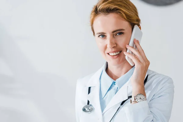 Medico felice guardando la fotocamera e sorridendo mentre parla su smartphone — Foto stock