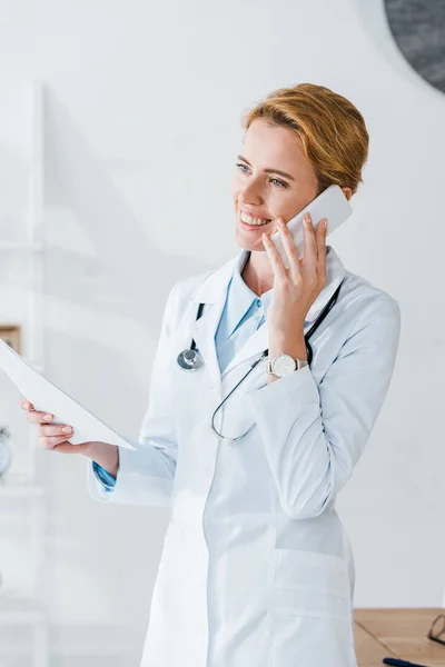 Feliz médico sosteniendo papel y sonriendo mientras habla en el teléfono inteligente - foto de stock