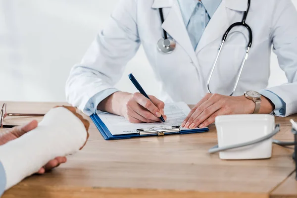 Vista recortada del paciente con el brazo roto cerca del médico escribiendo en el portapapeles - foto de stock