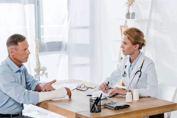 Atractivo médico mirando al hombre con el brazo roto mientras escribe el diagnóstico en la clínica - foto de stock