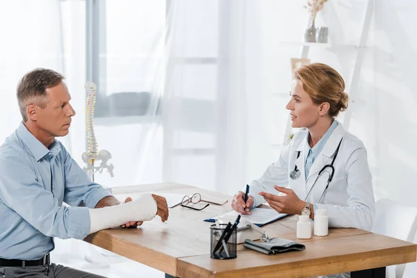 Attractive doctor looking at man with broken arm while holding pen in clinic — Stock Photo