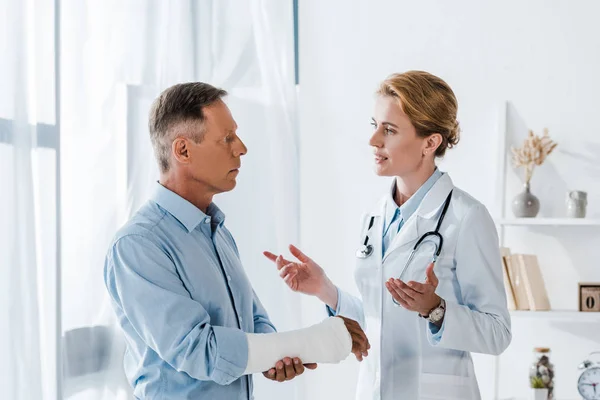 Attractive doctor gesturing near handsome man with broken arm in clinic — Stock Photo