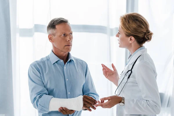 Привабливий лікар жестами біля людини з розбитим рукою в клініці — стокове фото