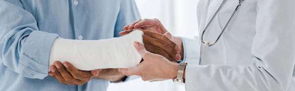 Tiro panorámico del médico tocando brazo roto del hombre en la clínica - foto de stock