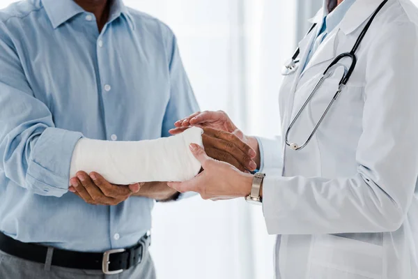 Vista recortada del médico tocando brazo roto en el hombre lesionado en la clínica - foto de stock