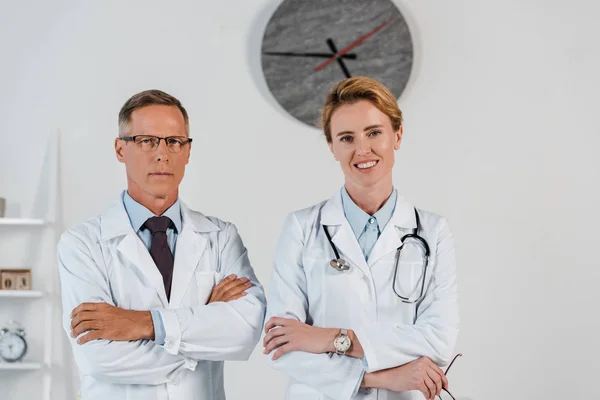Guapo médico de pie con los brazos cruzados cerca alegre colega en la clínica - foto de stock
