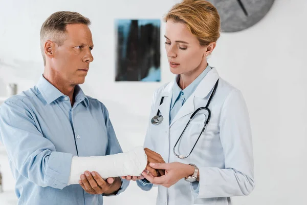 Привлекательный врач смотрит на сломанную руку на красивого мужчину в клинике — стоковое фото