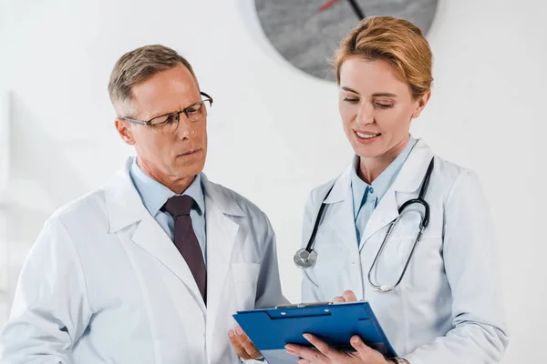 Guapo médico de pie cerca feliz compañero de trabajo y mirando portapapeles - foto de stock