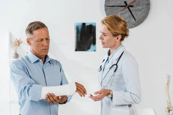 Attractive doctor looking at handsome man with broken arm — Stock Photo