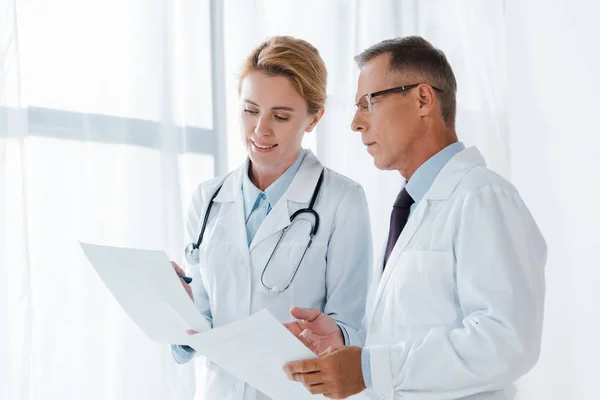 Cheerful doctor holding paper near handsome coworker in glasses — Stock Photo