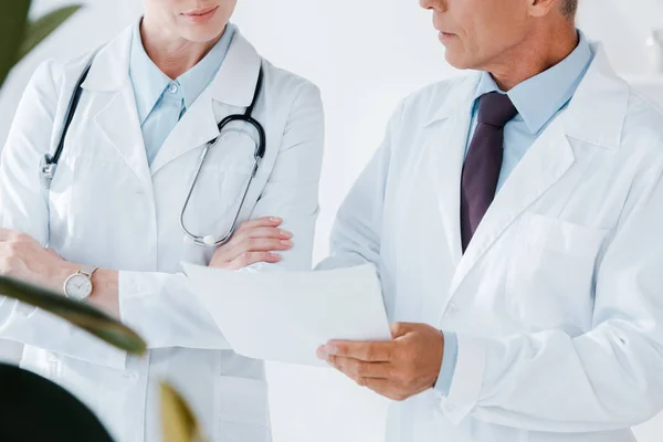 Vista ritagliata del medico in possesso di carta bianca vicino collega con braccia incrociate — Stock Photo