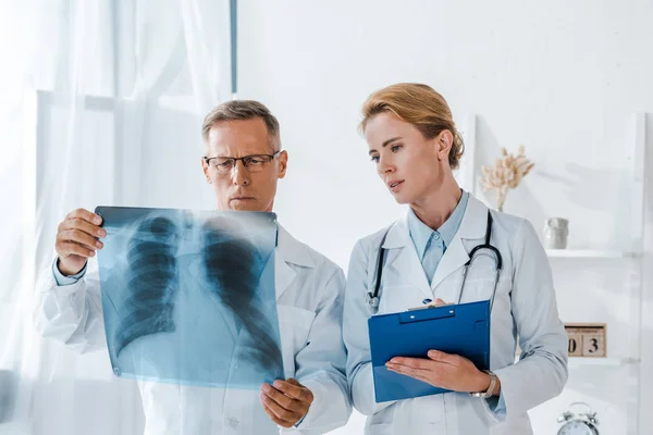 Atractivo médico sujetando portapapeles y mirando rayos X cerca de compañero de trabajo - foto de stock