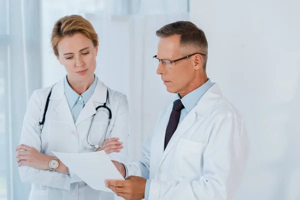 Man in glasses and white coat looking at blank paper near coworker with crossed arms — Stock Photo