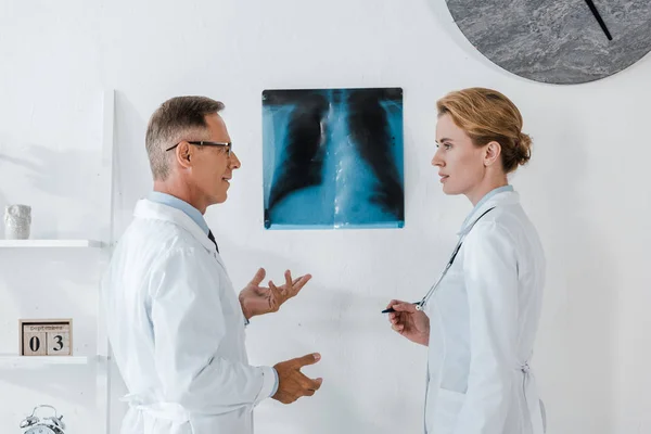 Лікар в окулярах жестикулює біля рентгенівського випромінювання, дивлячись на привабливого колеги — стокове фото