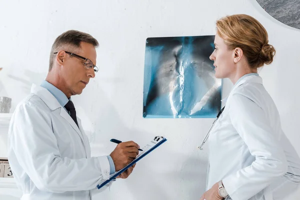 Hombre guapo en gafas de escritura mientras sostiene portapapeles cerca de rayos X y atractivo compañero de trabajo - foto de stock