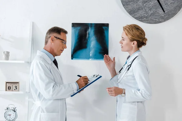 Schöner Mann mit Brille schreibt, während er Klemmbrett in der Nähe von Röntgen hält und attraktiver Kollege gestikuliert in Klinik — Stockfoto
