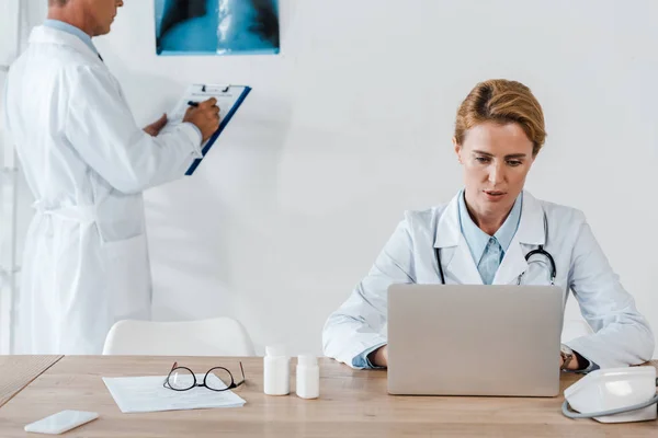Обрезанный вид врача, стоящего рядом с рентгеном и привлекательного коллеги с помощью ноутбука — стоковое фото
