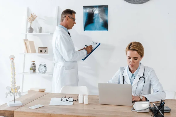 Beau médecin debout près de rayons X et collègue attrayant en utilisant un ordinateur portable — Photo de stock