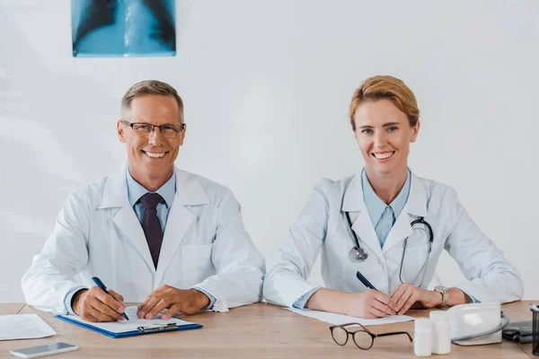 Cheerful doctors looking at camera and smiling while sitting near table — Stock Photo