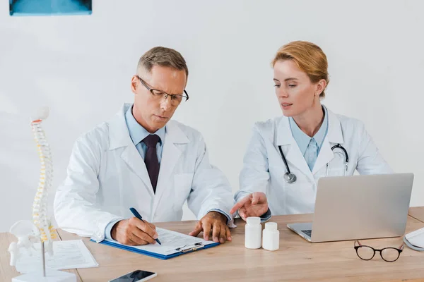 Atractivo médico señalando con el dedo cerca de la computadora portátil y compañero de trabajo en gafas de diagnóstico de escritura - foto de stock