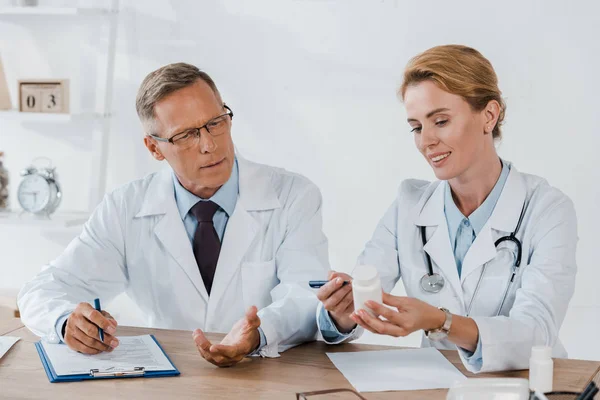 Médico bonito em óculos gesticulando enquanto olha para garrafa perto colega alegre — Fotografia de Stock