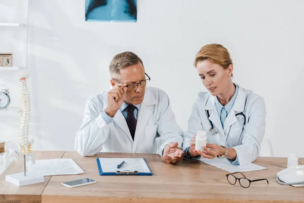 Arzt berührt Brille beim Flaschenanblick in der Nähe attraktiver Mitarbeiter — Stockfoto