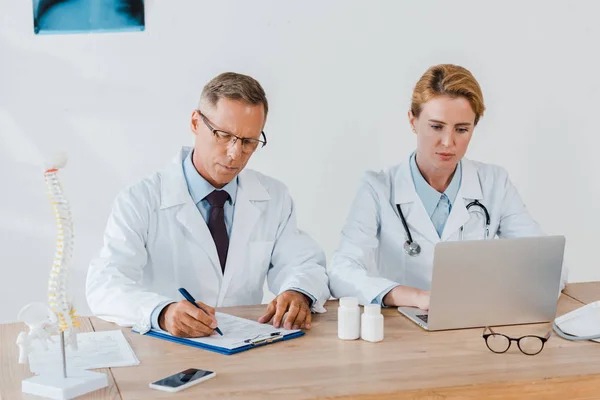 Doctor writing diagnosis near coworker using laptop in hospital — Stock Photo