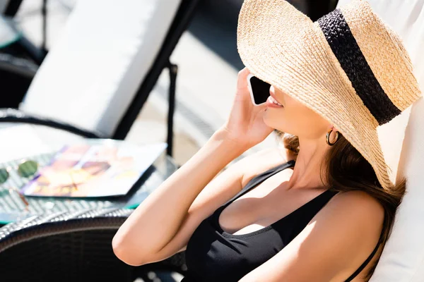 Девушка в купальнике и соломенной шляпе лежит на шезлонге и разговаривает на смартфоне на курорте — стоковое фото