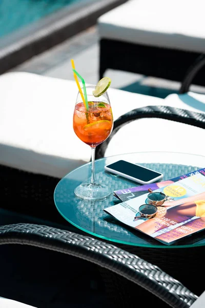 Стол с коктейлем, сигаретами, смартфоном и солнцезащитными очками рядом с шезлонгом на курорте — стоковое фото