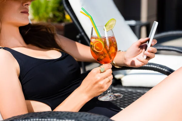 Vue recadrée de la fille en maillot de bain avec cocktail sur chaise longue en utilisant smartphone — Photo de stock