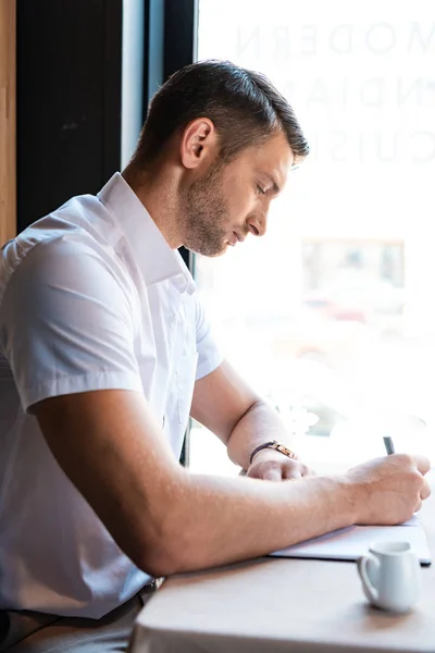 Vista laterale di focalizzato bell'uomo che scrive nel libro di testo nel caffè — Foto stock