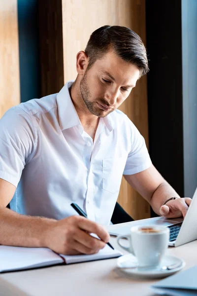 Focused handsome freelancer writing in textbook and typing on laptop keyboard in cafe — Stock Photo