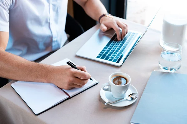 Visão parcial da escrita freelancer no livro didático e digitação no teclado do laptop no café — Fotografia de Stock