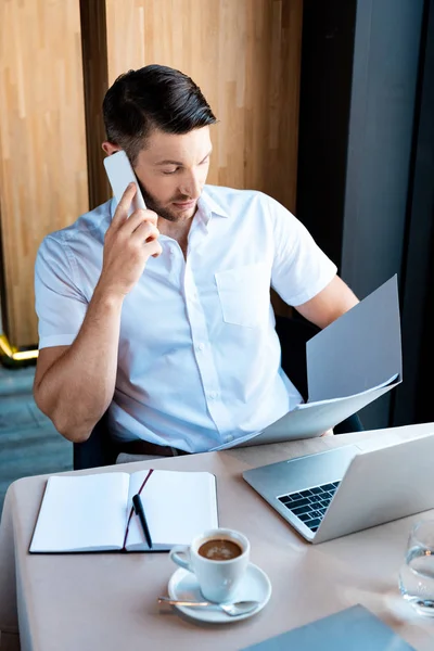 Hombre sosteniendo la carpeta y hablando en el teléfono inteligente mientras está sentado en la cafetería - foto de stock