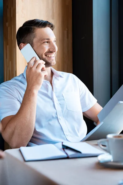 Sonriente hombre sosteniendo la carpeta y hablando en el teléfono inteligente mientras está sentado en la cafetería - foto de stock
