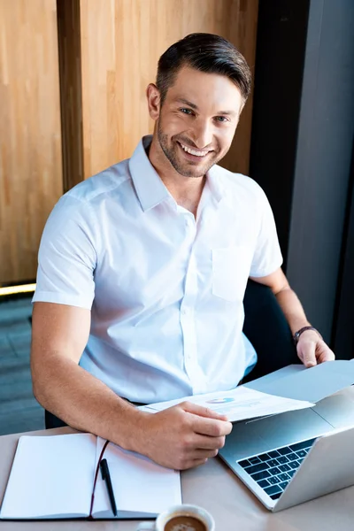 Sonriente hombre sosteniendo carpeta mientras está sentado en la mesa con el ordenador portátil en la cafetería - foto de stock