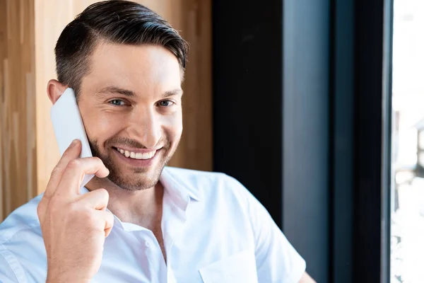 Усміхнений чоловік у білій сорочці розмовляє на смартфоні і дивиться на камеру — стокове фото