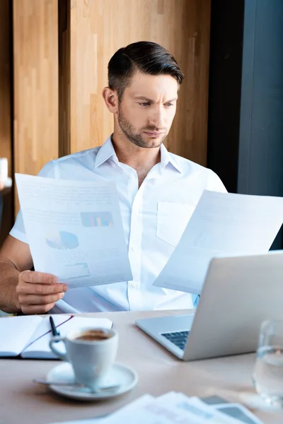 Verwirrter Mann sitzt mit Laptop am Tisch und schaut sich Dokumente im Café an — Stockfoto