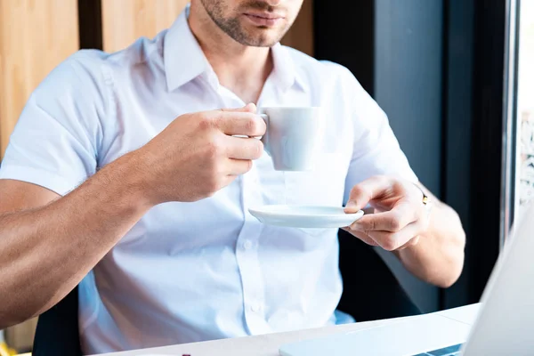 Vista recortada del hombre sosteniendo platillo y taza de café en la cafetería - foto de stock
