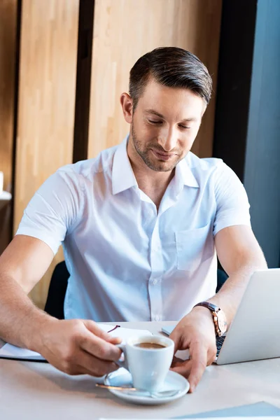 Souriant freelance tenant soucoupe et tasse de café tout en étant assis à table avec ordinateur portable dans le café — Photo de stock