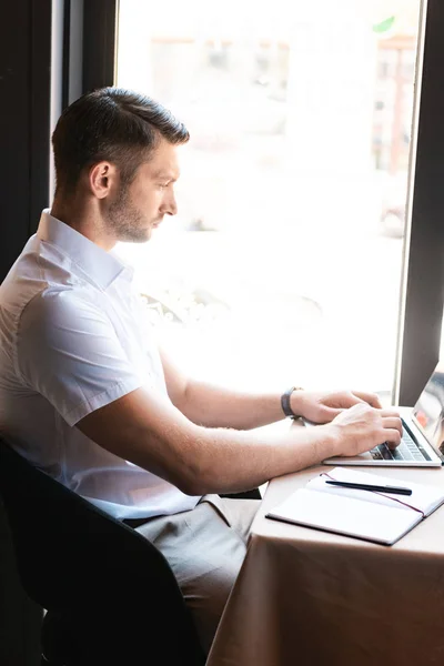 Side view of focused freelancer typing on laptop keyboard while sitting in cafe — Stock Photo