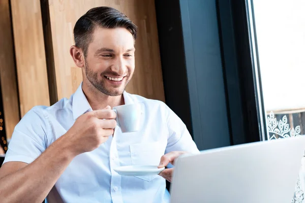 Sonriente freelancer sosteniendo platillo y taza de café mientras mira sceeen portátil - foto de stock