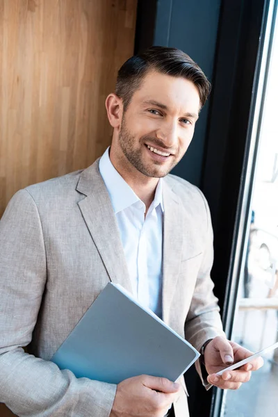 Smiling businessman holding folder and smartphone and looking at camera — Stock Photo
