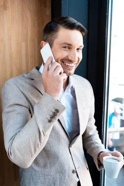 Sonriente hombre de negocios sosteniendo una taza de café y hablando en el teléfono inteligente en la cafetería - foto de stock