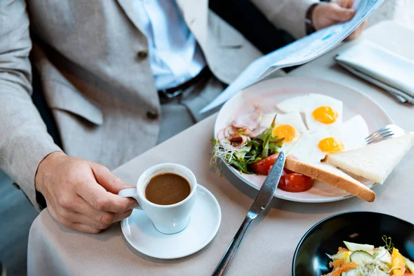 Cropped view of businessman holding cup of coffee and reading newspaper in cafe — Stock Photo