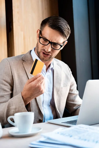 Confused businessman in glasses with credit card and laptop in cafe — Stock Photo
