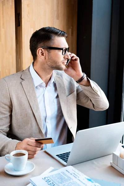 Homem de negócios em óculos com cartão de crédito e laptop conversando no smartphone no café — Fotografia de Stock